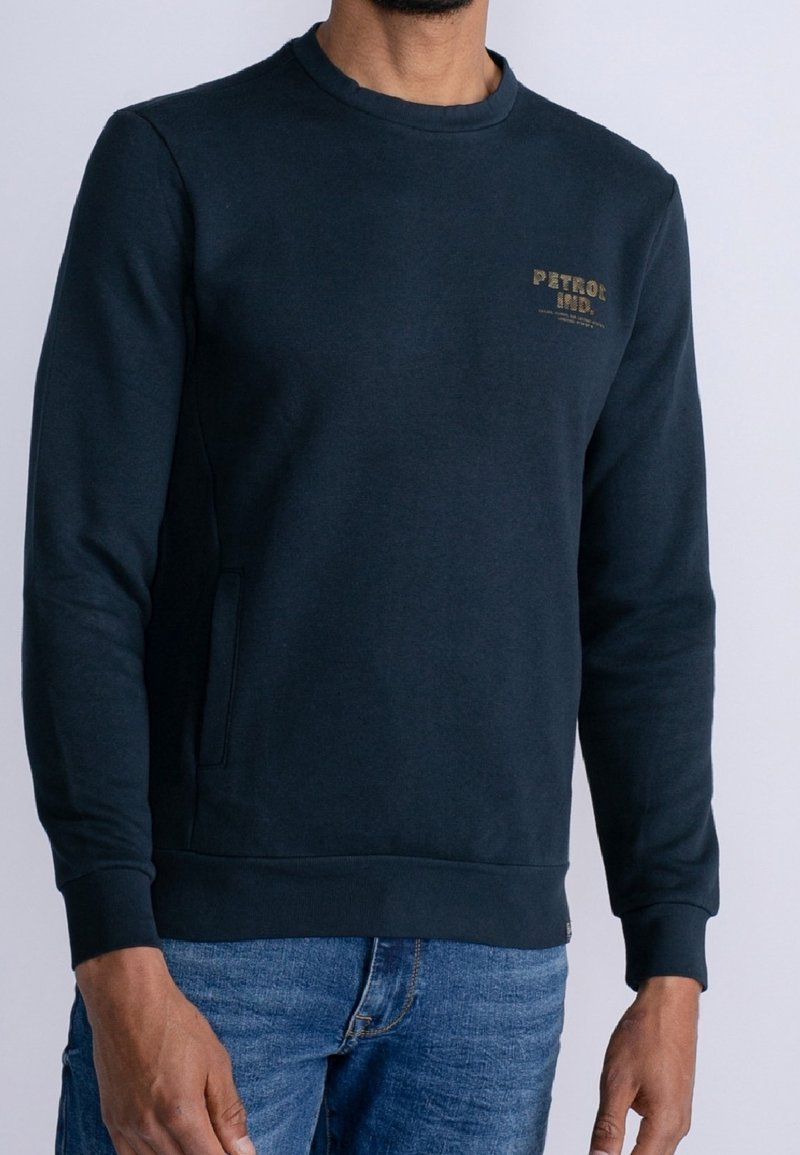 Sudadera azul oscuro con cuello redondo y mangas largas. Presenta un bolsillo en la parte frontal y un logo de texto dorado en el pecho. Textura de tela suave.