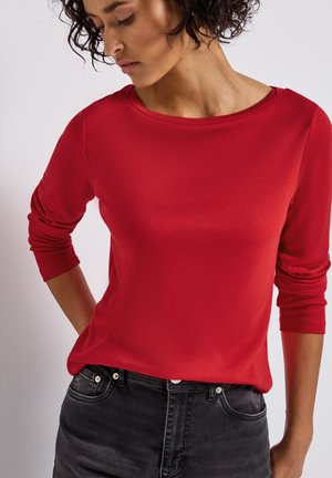 Maglietta rossa a maniche lunghe con scollatura ampia, caratterizzata da una texture liscia. Abbinata a jeans grigio scuro a vita alta, che mostrano linee pulite e dettagli minimi.