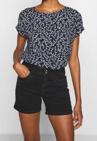 Top blu navy a maniche corte con un motivo floreale bianco, abbinato a pantaloni corti neri a vita alta. Il top è morbido e realizzato in tessuto leggero.