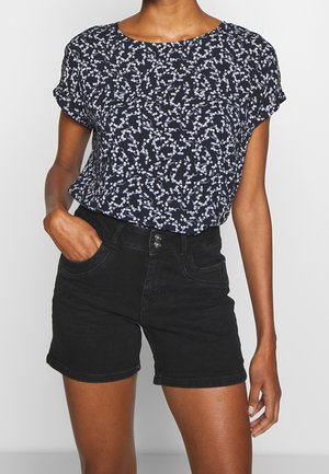 Mørkeblå kortærmet bluse med et hvidt blomsterdesign, parret med højhalsede sorte shorts. Blusen er løs og lavet af let materiale.