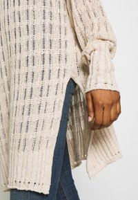 Pull en tricot beige avec un motif texturé et lâche. Présente des poignets côtelés et des fentes latérales, associé à un jean bleu foncé.