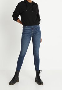 Sudadera cortada negra con cordones, combinada con jeans ajustados azules y botas de combate negras con cordones y suela gruesa.