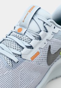 Šviesiai mėlyna sportinė batai su tinkliniais viršutiniais, atspindinčiu pilku Nike logotipu, tekstūruotomis raištelėmis ir oranžinėmis akcentais ant liežuvėlio bei vidurinės dalies.