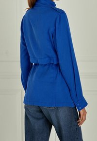 Woman wearing a bright blue long-sleeve jacket with buttoned cuffs and high collar, paired with dark blue denim jeans, shown from the back.