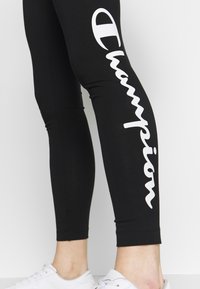 Czarne legginsy z elastycznego materiału z białym logo "Champion" nadrukowanym pionowo wzdłuż lewej nogawki. Dolna krawędź jest dopasowana i gładka.