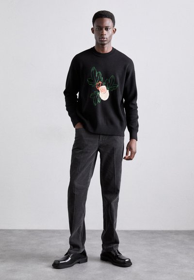 Black sweater with floral embroidery in green and beige, paired with dark gray pants and black shoes, creating a monochromatic outfit.