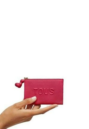 Cartera de cuero rosa con el logo grabado "TOUS", cierre de cremallera y tirador en forma de corazón. Textura suave, forma rectangular, diseño compacto.