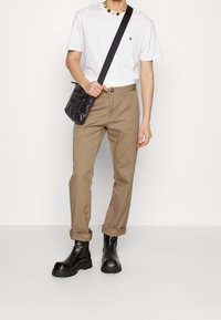 Tan katoenen broek met opgerolde pijpen, gecombineerd met een witte t-shirt en zwarte glanzende enkellaarsjes. Accessoires omvatten een zwarte gewatteerde tas.