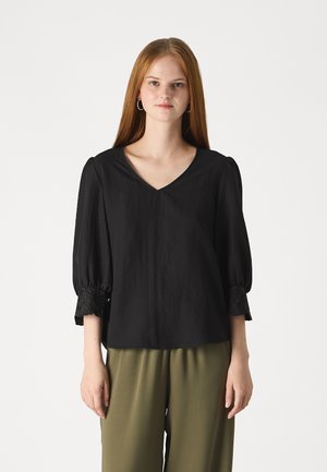 Black blouse with a V-neckline and short puff sleeves, featuring embroidered lace accents at the cuffs. Paired with olive green pants.