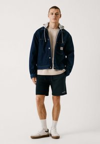 Chaqueta de mezclilla con forro de sudadera gris, camiseta beige, pantalones cortos oscuros, zapatillas blancas con suelas marrones, de pie sobre un fondo claro.