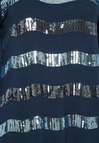 Top blu navy con strisce orizzontali di paillettes in diverse tonalità di blu e argento, creando un motivo ondulato su uno sfondo solido.