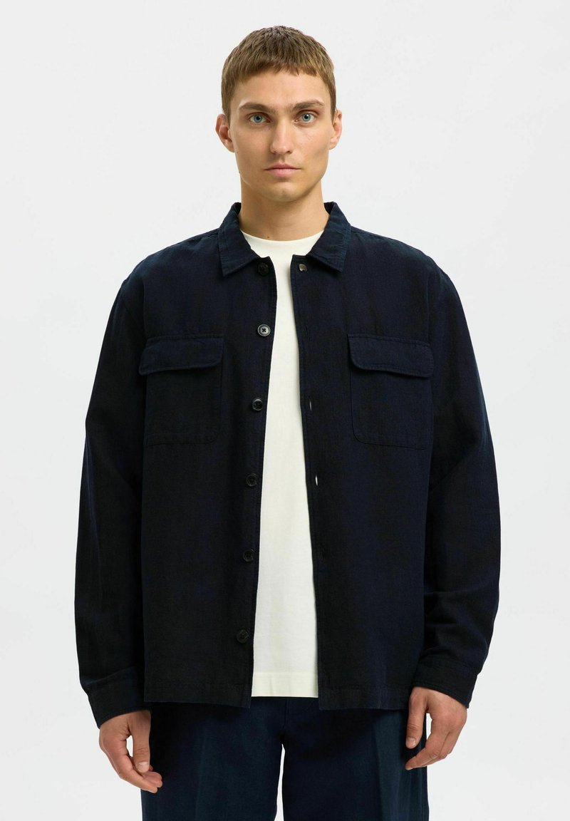 Marineblauwe katoenen jas met twee opgestikte zakken, knoopsluiting, kraag en lange mouwen. Gelaagd boven een wit T-shirt.