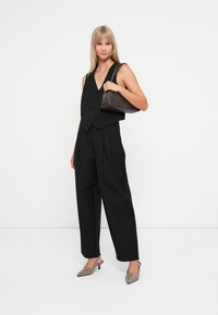 Gilet nero con scollatura a forma di U, abbinato a pantaloni neri a vita alta. Il modello tiene in mano una borsa in pelle marrone scuro e indossa tacchi grigi.