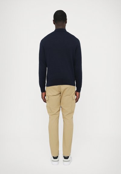 Tengerészkék pulóver texturált kivitelben, bézs színű cargo nadrág oldalzsebekkel, valamint fehér sportcipő fekete részletekkel.