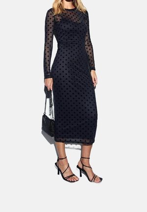 Kobieta ubrana w czarną, przejrzystą sukienkę midi z długimi rękawami i czarnymi kropkami, trzymająca czarną torebkę na łańcuszku i nosząca czarne sandały na obcasie z paskami.