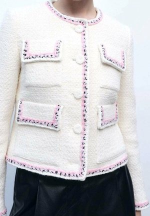 Veste blanche texturée avec bordure rose et noire, dotée de quatre poches à rabat devant et de boutons ronds, portée avec un bas noir.