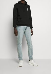 Svart huvtröja med grafik på bröstet, kombinerad med ljusblå jeans och vita sneakers. Huvtröjan har en känguruficka och dragsnören.