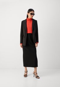 Black blazer over a red ribbed turtleneck, black midi skirt, and black strappy heels, with dark sunglasses. Smooth fabric and tailored fit.