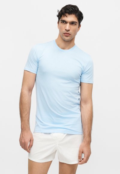 Νέος άνδρας που φοράει ανοιχτό μπλε εφαρμοστή κοντομάνικη πουκάμισα και λευκά σορτς στέκεται μπροστά σε απλό ανοιχτόχρωμο φόντο.