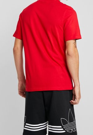 Rood katoenen T-shirt met korte mouwen, effen ontwerp, gezien vanaf de achterkant, gedragen met zwarte korte broek met witte gestreepte accenten en logo.