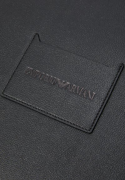 Zwarte textuur leren label genaaid op een zwart leren oppervlak met het reliëf van het Emporio Armani-logo en de adelaar in het midden.