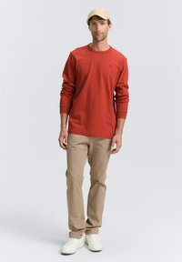 Langärmliges rotes Baumwoll-T-Shirt, kombiniert mit hellen Khakihosen und weißen Turnschuhen. Eine beigen Mütze rundet den Look ab. Einfaches Design ohne Muster.