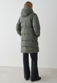 Manteau long et rembourré en vert olive avec une grande capuche ajustée, des coutures verticales et une taille cintrée. Associé à un jean foncé et des chaussures noires.