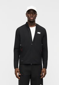 Άνδρας που φοράει μαύρο μπουφάν με φερμουάρ και παντελόνι με λευκά λογότυπα, λευκή μπλούζα και λευκό καπέλο, στέκεται μπροστά σε απλό γκρι φόντο.