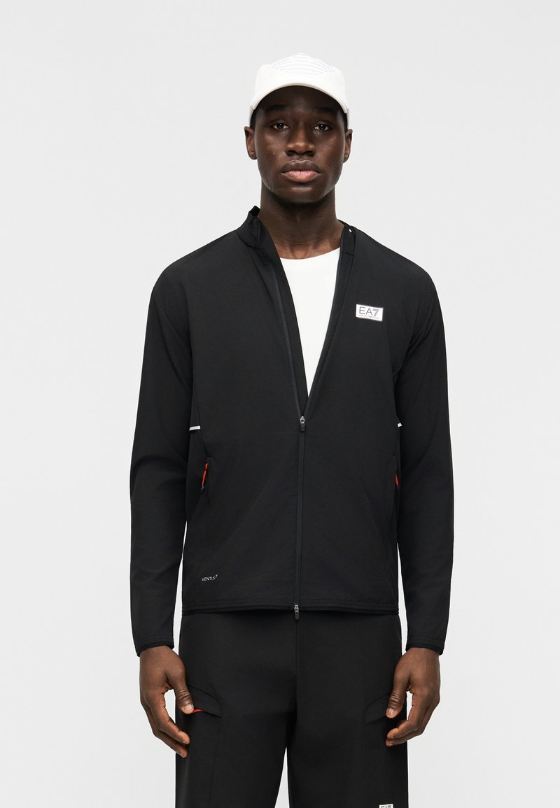 Άνδρας που φοράει μαύρο μπουφάν με φερμουάρ και παντελόνι με λευκά λογότυπα, λευκή μπλούζα και λευκό καπέλο, στέκεται μπροστά σε απλό γκρι φόντο.