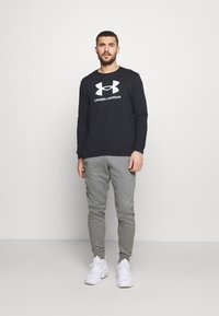 Czarna koszula z długim rękawem z białym logo "Under Armour", w połączeniu z szarymi dresami. Białe buty sportowe dopełniają strój.