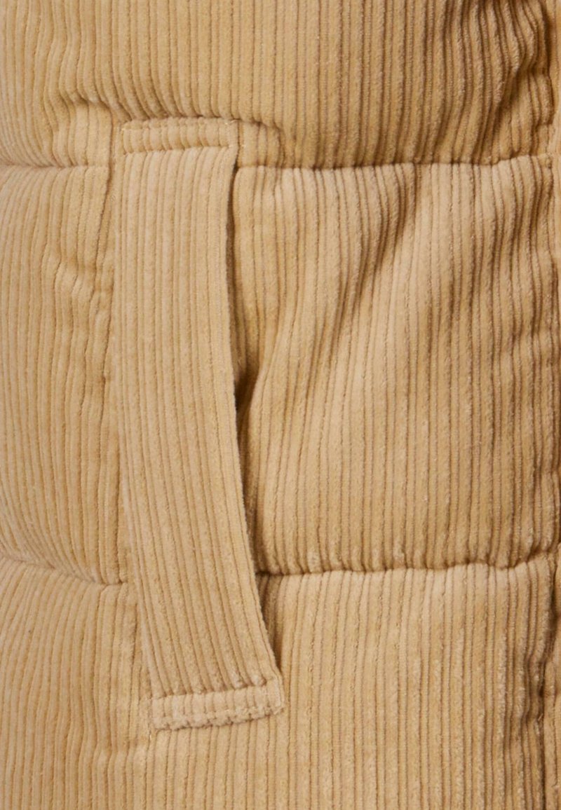 Material de pana beige con acanalado vertical, que cuenta con un bolsillo lateral. La textura es suave y esponjosa, lo que aumenta la calidez y la comodidad.