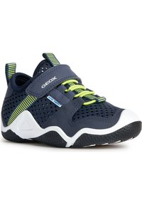 Zapatillas infantiles de color azul marino, hechas de malla transpirable, con una correa de velcro, detalles verdes y una suela de goma negra texturizada.