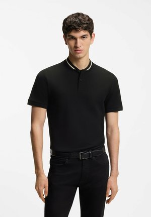 Junger Mann trägt ein schwarzes Polohemd mit weiß gestreiftem Kragen, schwarzen Gürtel und schwarze Hose, stehend vor einem schlichten weißen Hintergrund.