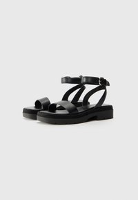 Sandales en cuir noir avec une semelle plate, présentant deux larges sangles sur le pied et une sangle au niveau de la cheville avec une boucle en métal. Surface texturée.
