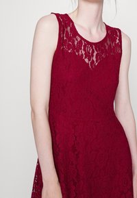 Burgundy klänning med ett blommönstrat spetsdesign, ärmlös, med en genomskinlig spetskrage och slät tygtextur.