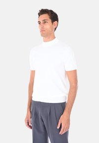 Uomo con capelli ricci corti che indossa una camicia bianca aderente a maniche corte e pantaloni grigi larghi a pieghe, in piedi su uno sfondo bianco.