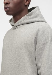 Gros plan sur une personne portant un sweat à capuche gris clair, montrant le côté du visage et le haut du torse.