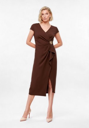 Ξανθιά γυναίκα που φοράει καφέ φόρεμα τύπου τούλι μήκους μέχρι το γόνατο με διακοσμητικό καρφίτσα και μπεζ ψηλοτάκουνα, στέκεται με τα χέρια πίσω από την πλάτη σε λευκό φόντο.