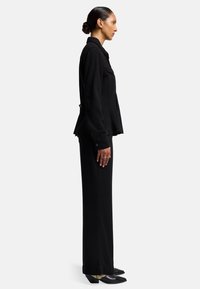 Giacca nera con texture e colletto, abbinata a pantaloni neri su misura. Le scarpe hanno un design con punta affilata e tacco quadrato.