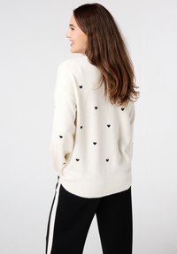 Romantische crème trui met zwarte hartpatronen, ronde halslijn en lange mouwen, gecombineerd met zwarte broeken met zijsstripes. Zachte textuur.