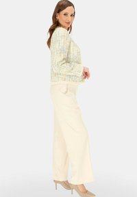 Kobieta stojąca bokiem, ubrana w jasną pastelową kurtkę z tweedu, kremowe spodnie o szerokich nogawkach oraz beżowe wysokie obcasy na jednolitym tle.