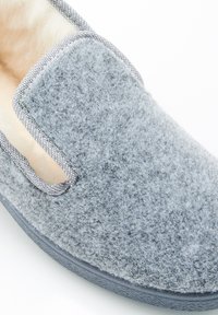 Pantofole in feltro grigio con forma arrotondata, lati aperti e un bordo testurizzato a contrasto. L'interno morbido offre comfort.