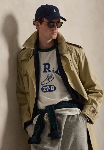 Άνδρας που φοράει ναυτικό καπέλο, γυαλιά ηλίου, μπεζ καμπαρντίνα, λευκή μπλούζα με σχέδιο, γκρι φόρμα και καρό πουκάμισο δεμένο γύρω από τη μέση, ποζάροντας δίπλα σε τοίχο.