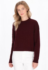 Burgunderfarbener Kabelstrickpullover mit rundem Ausschnitt und langen Ärmeln, mit einem gerippten Saum. Kombiniert mit weißen Hosen.