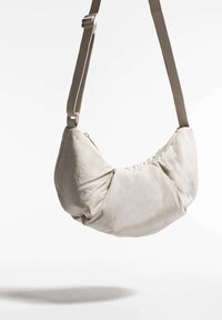 Beige szövet válltáska, puha, lekerekített formával, seamless design-nal és állítható beige pánttal. Cipzáras záródás részlete látható.