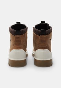 Botas de cuero marrón hasta el tobillo con un cuello de fieltro gris, suela de goma blanca y una tira de agarre negra en la parte posterior. Presentan costuras duraderas y un acabado texturizado.