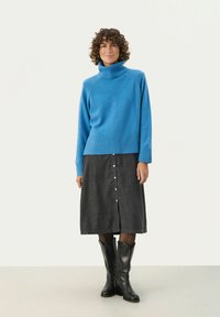 Modra pulover turtleneck z rebrastim robom, v kombinaciji s temno jeans krilom, ki ima bele gumbe, in črnimi kavbojkami do kolen.