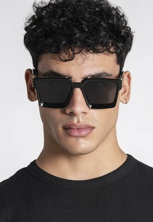 Jeune homme aux cheveux noirs bouclés portant de grandes lunettes de soleil rectangulaires noires et un t-shirt noir à col rond côtelé, fond neutre.