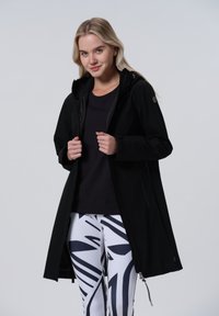 Veste noire d'extérieur avec capuche, présentant une texture lisse et des poches zippées, associée à des leggings à motifs blancs et noirs.