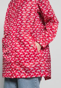 Czerwona peleryna przeciwdeszczowa z powtarzającym się białym wzorem psów, z bocznymi kieszeniami i elastycznymi mankietami, wykonana z gładkiego, lekkiego materiału.